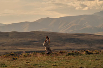 "Pregnant woman standing in a grassy field at sunset, surrounded by wildflowers, with rolling hills and mountains in the background under a cloudy sky." "Pregnant woman standing in a grassy field at sunset, surrounded by wildflowers, with rolling hills and mountains in the background under a cloudy sky."